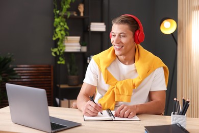 Photo of Man in headphones taking notes while passing online test via laptop at table indoors