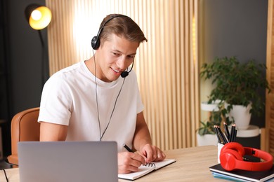 Photo of Man in headset taking notes while passing online test via laptop at table indoors