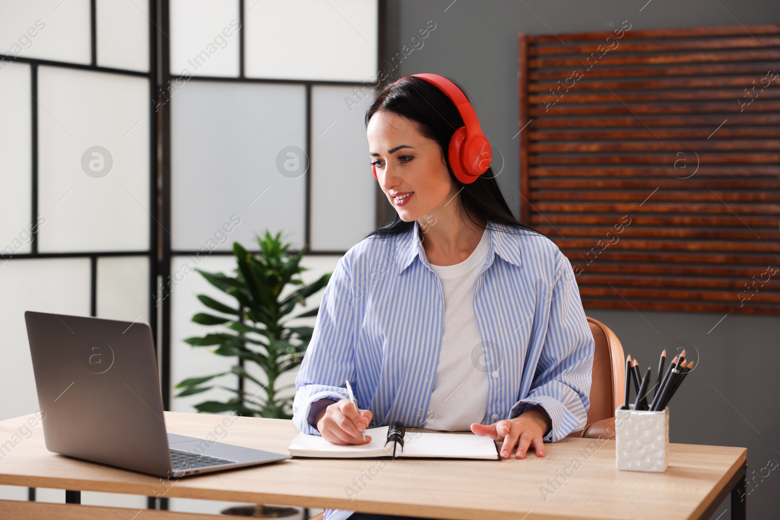 Photo of Woman in headphones taking notes while passing online test via laptop at table indoors