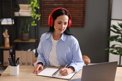 Photo of Woman in headphones taking notes while passing online test via laptop at table indoors