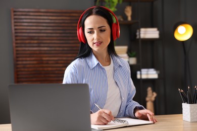 Photo of Woman in headphones taking notes while passing online test via laptop at table indoors