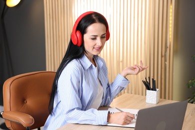 Photo of Woman in headphones taking notes while passing online test via laptop at table indoors