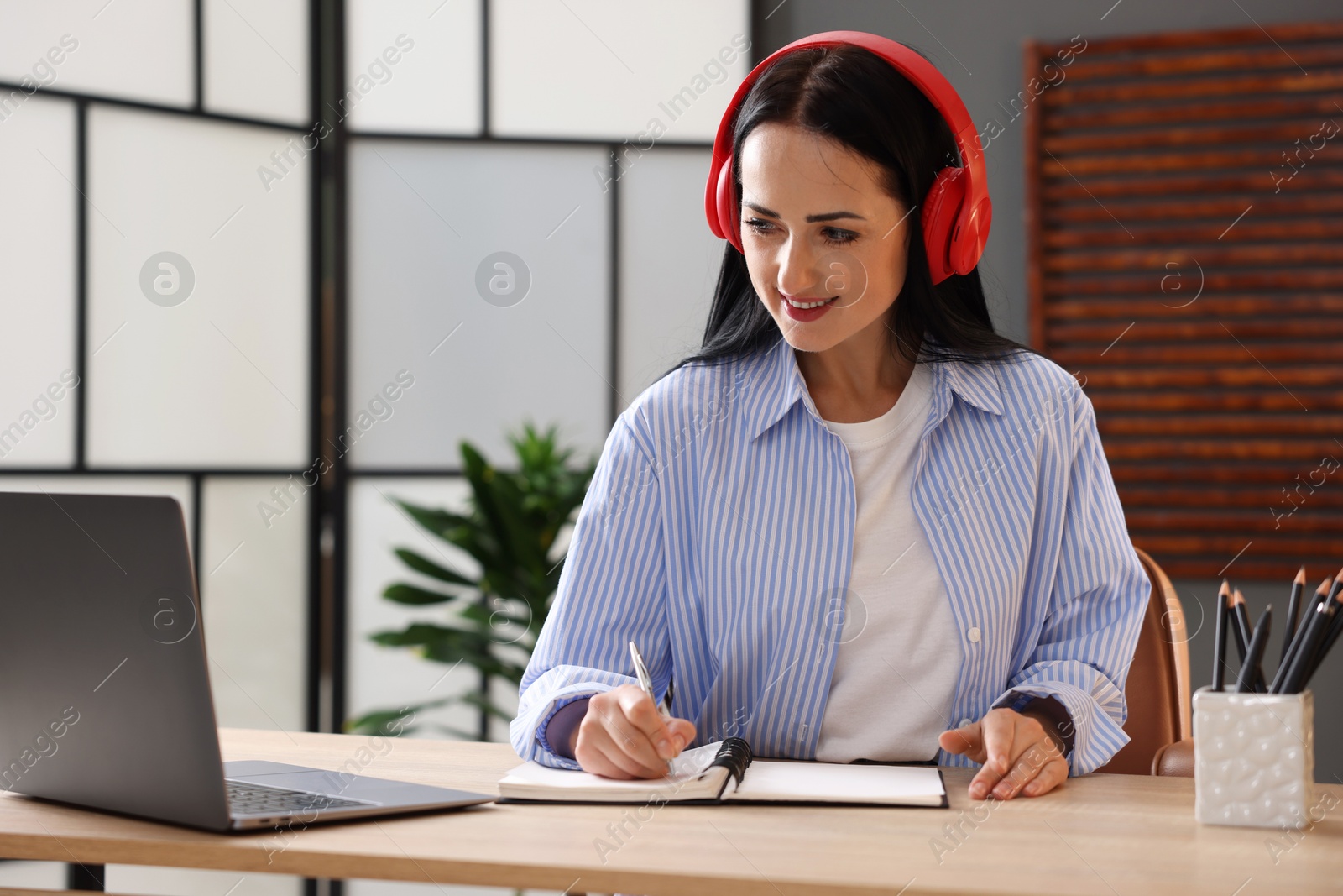 Photo of Woman in headphones taking notes while passing online test via laptop at table indoors