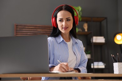 Photo of Woman in headphones taking notes while passing online test via laptop at table indoors