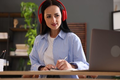 Photo of Woman in headphones taking notes while passing online test via laptop at table indoors