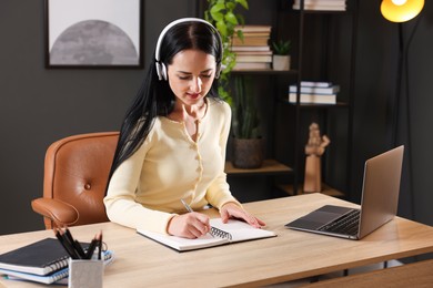 Photo of Woman in headphones taking notes while passing online test via laptop at table indoors