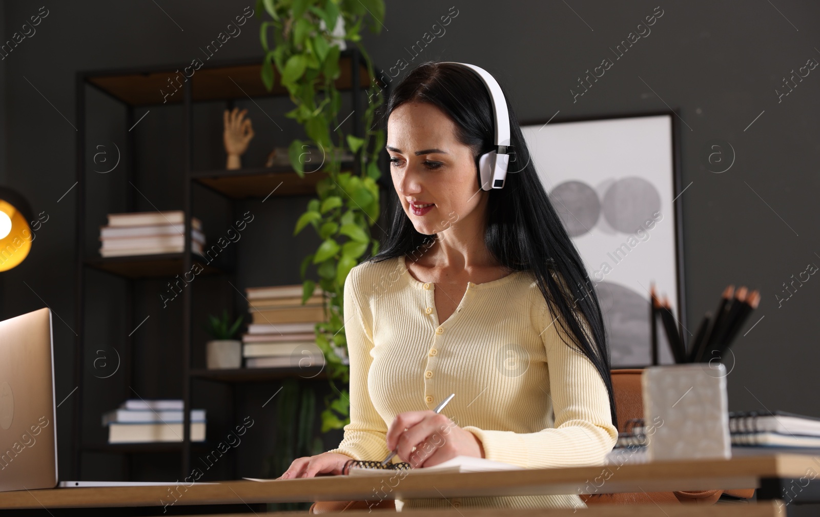 Photo of Woman in headphones taking notes while passing online test via laptop at table indoors