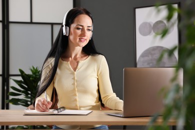 Photo of Woman in headphones taking notes while passing online test via laptop at table indoors