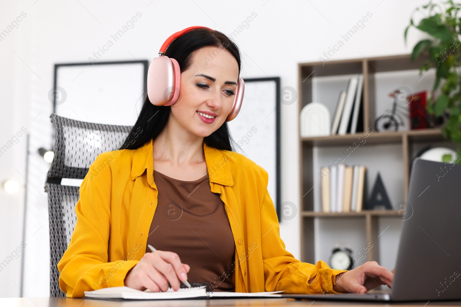 Photo of Woman in headphones taking notes while passing online test via laptop at table indoors
