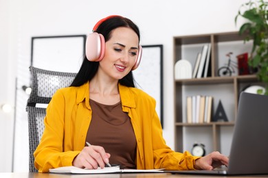 Photo of Woman in headphones taking notes while passing online test via laptop at table indoors