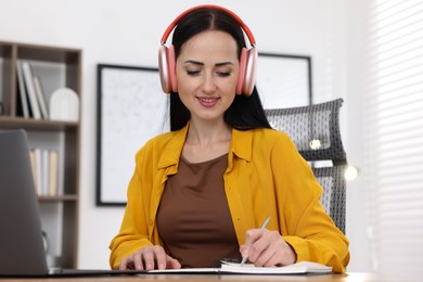 Photo of Woman in headphones taking notes while passing online test via laptop at table indoors