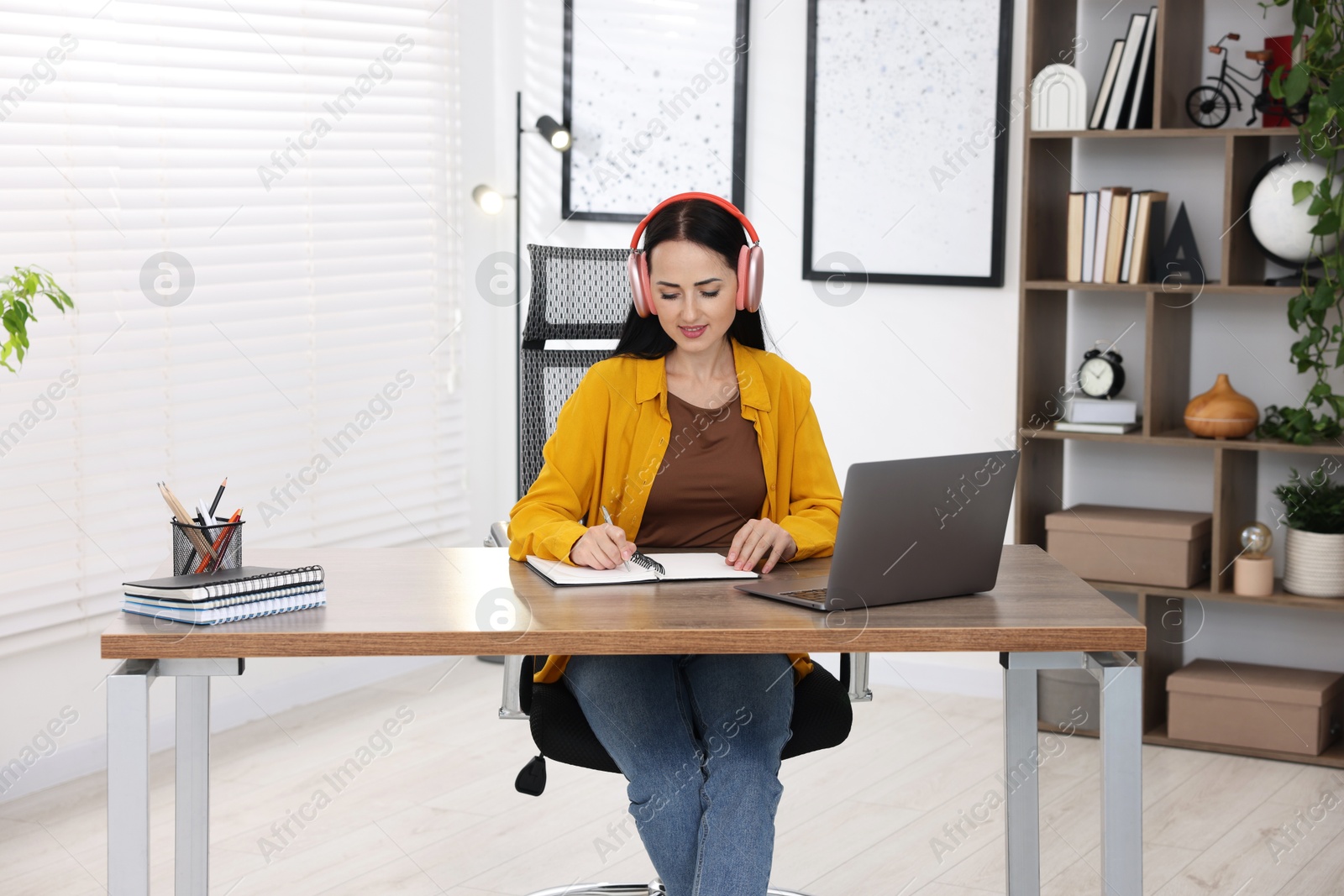 Photo of Woman in headphones taking notes while passing online test via laptop at table indoors
