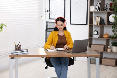 Photo of Woman in headphones taking notes while passing online test via laptop at table indoors