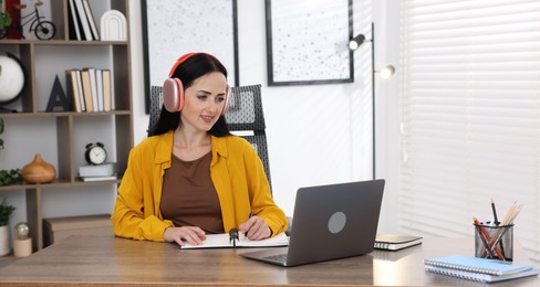 Photo of Woman in headphones taking notes while passing online test via laptop at table indoors