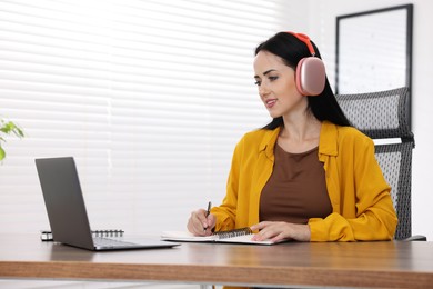 Photo of Woman in headphones taking notes while passing online test via laptop at table indoors