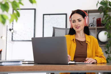 Photo of Woman in headphones taking notes while passing online test via laptop at table indoors