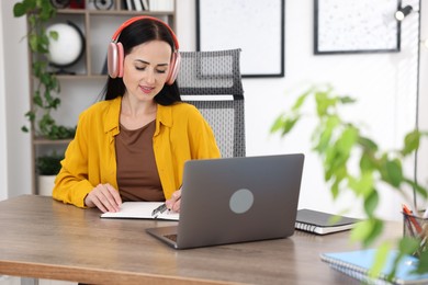 Photo of Woman in headphones taking notes while passing online test via laptop at table indoors