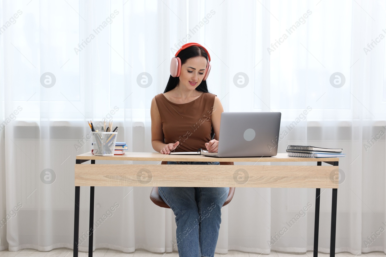 Photo of Woman in headphones taking notes while passing online test via laptop at table indoors