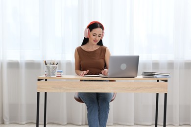 Photo of Woman in headphones taking notes while passing online test via laptop at table indoors