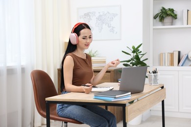 Photo of Woman in headphones passing online test via laptop at table indoors