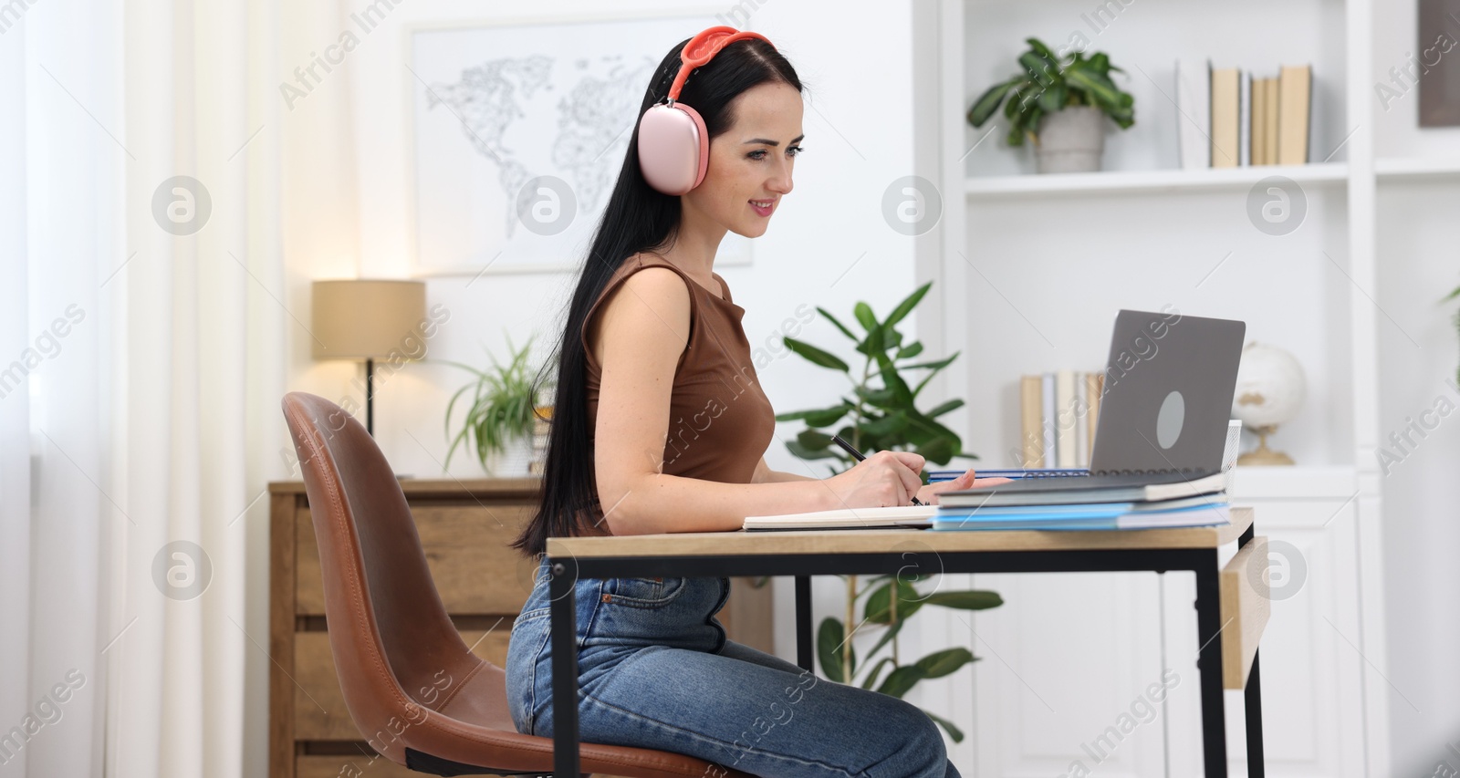 Photo of Woman in headphones taking notes while passing online test via laptop at table indoors