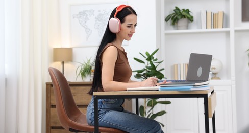Photo of Woman in headphones taking notes while passing online test via laptop at table indoors