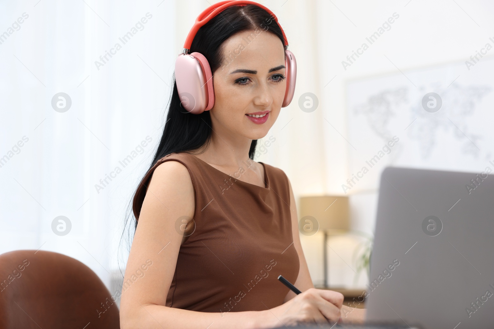 Photo of Woman in headphones taking notes while passing online test via laptop indoors