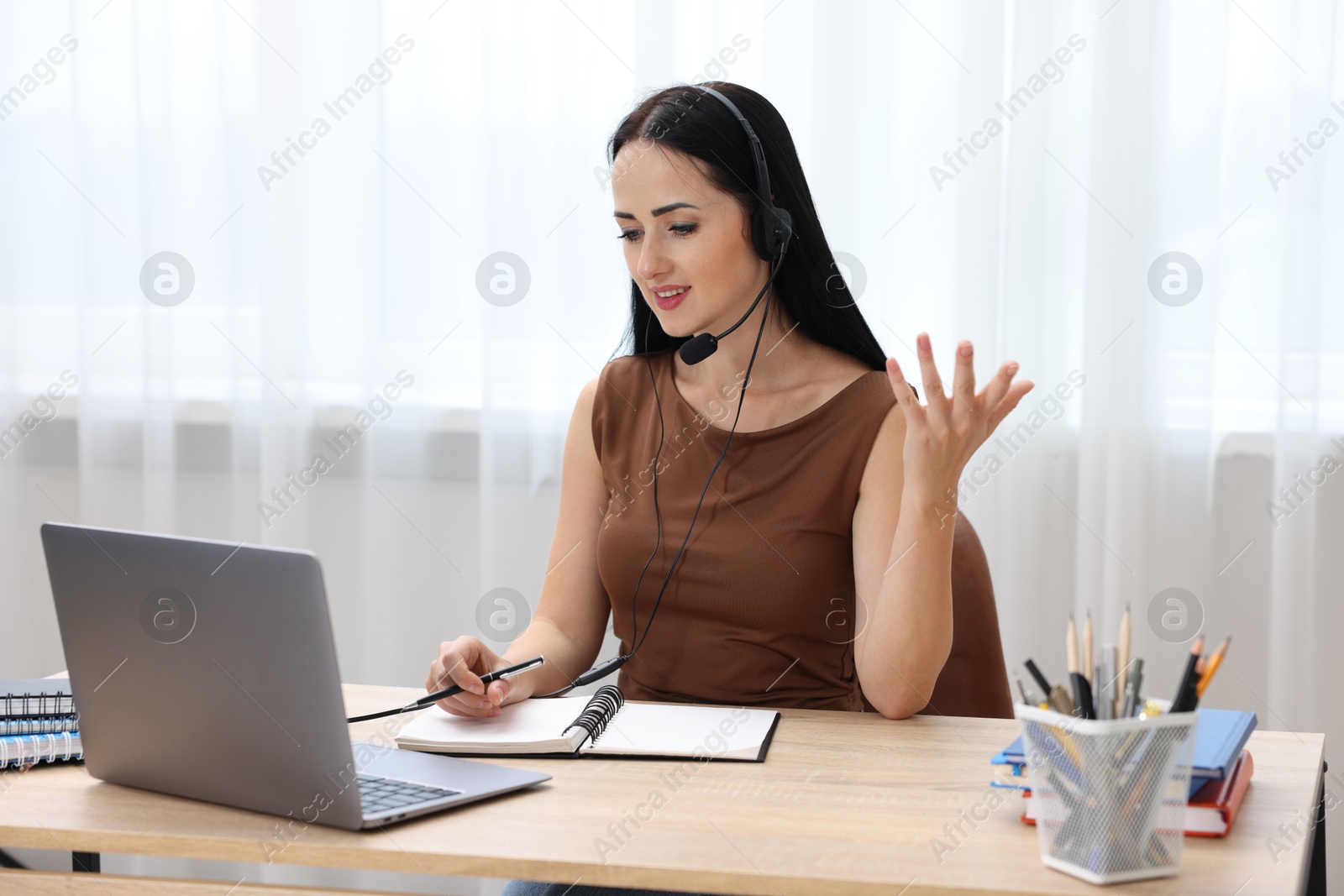 Photo of Woman in headset taking notes while passing online test via laptop at table indoors
