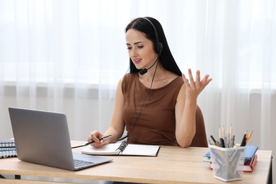 Photo of Woman in headset taking notes while passing online test via laptop at table indoors