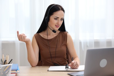 Photo of Woman in headset taking notes while passing online test via laptop at table indoors