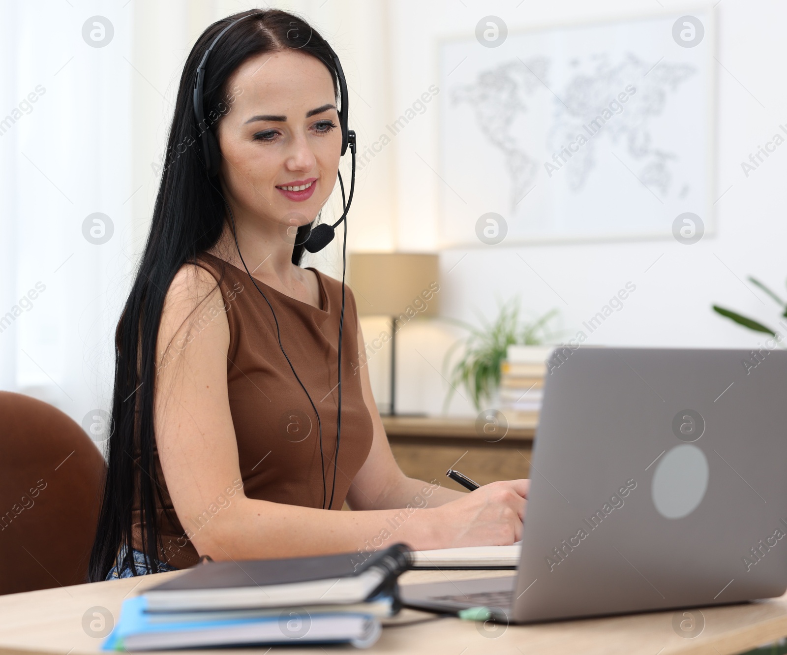Photo of Woman in headset taking notes while passing online test via laptop at table indoors