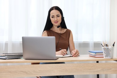 Photo of Woman in headset taking notes while passing online test via laptop at table indoors
