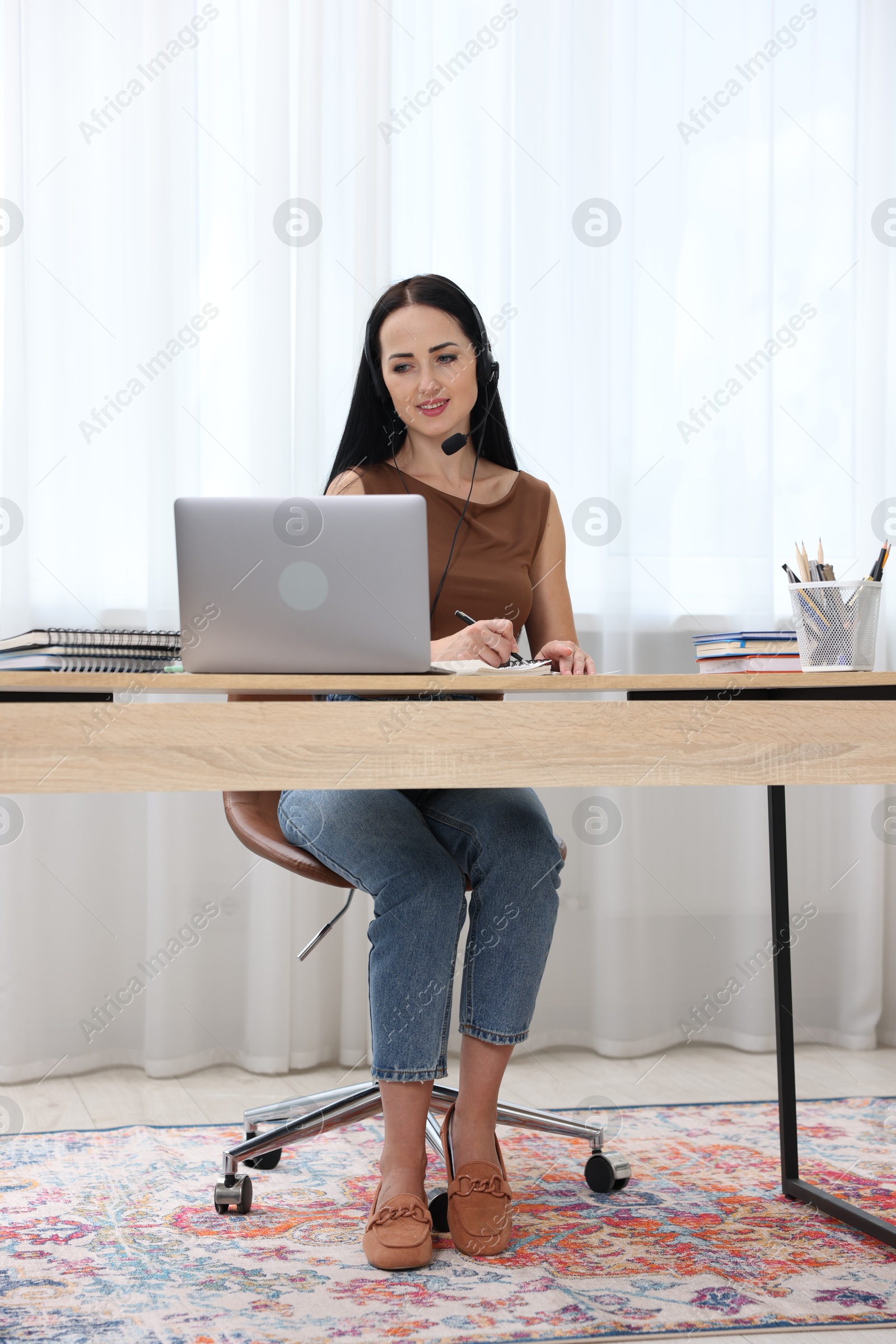 Woman in headset taking notes while passing online test via laptop at table indoors Photo of Woman in headset taking notes while passing online test via laptop at table indoors