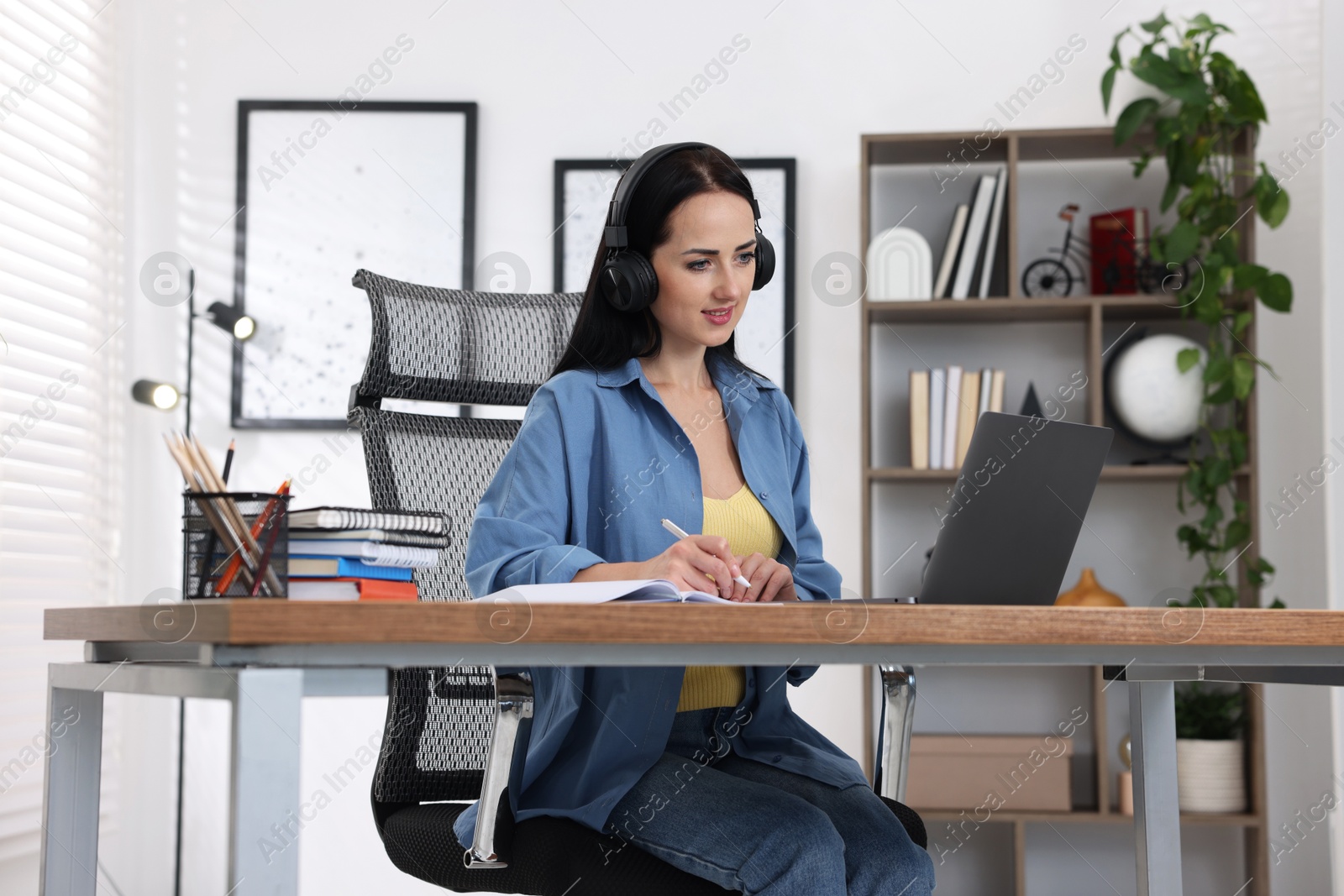 Woman in headphones taking notes while passing online test on laptop at table indoors Photo of Woman in headphones taking notes while passing online test on laptop at table indoors