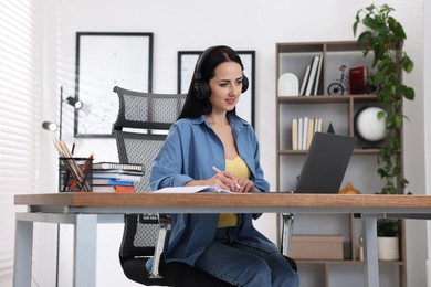 Photo of Woman in headphones taking notes while passing online test on laptop at table indoors