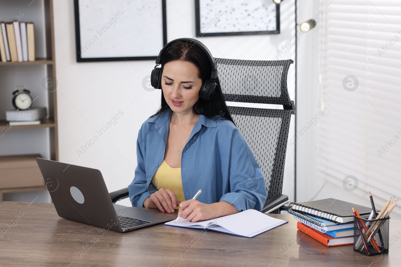 Woman in headphones taking notes while passing online test on laptop at table indoors Photo of Woman in headphones taking notes while passing online test on laptop at table indoors