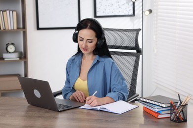 Photo of Woman in headphones taking notes while passing online test on laptop at table indoors
