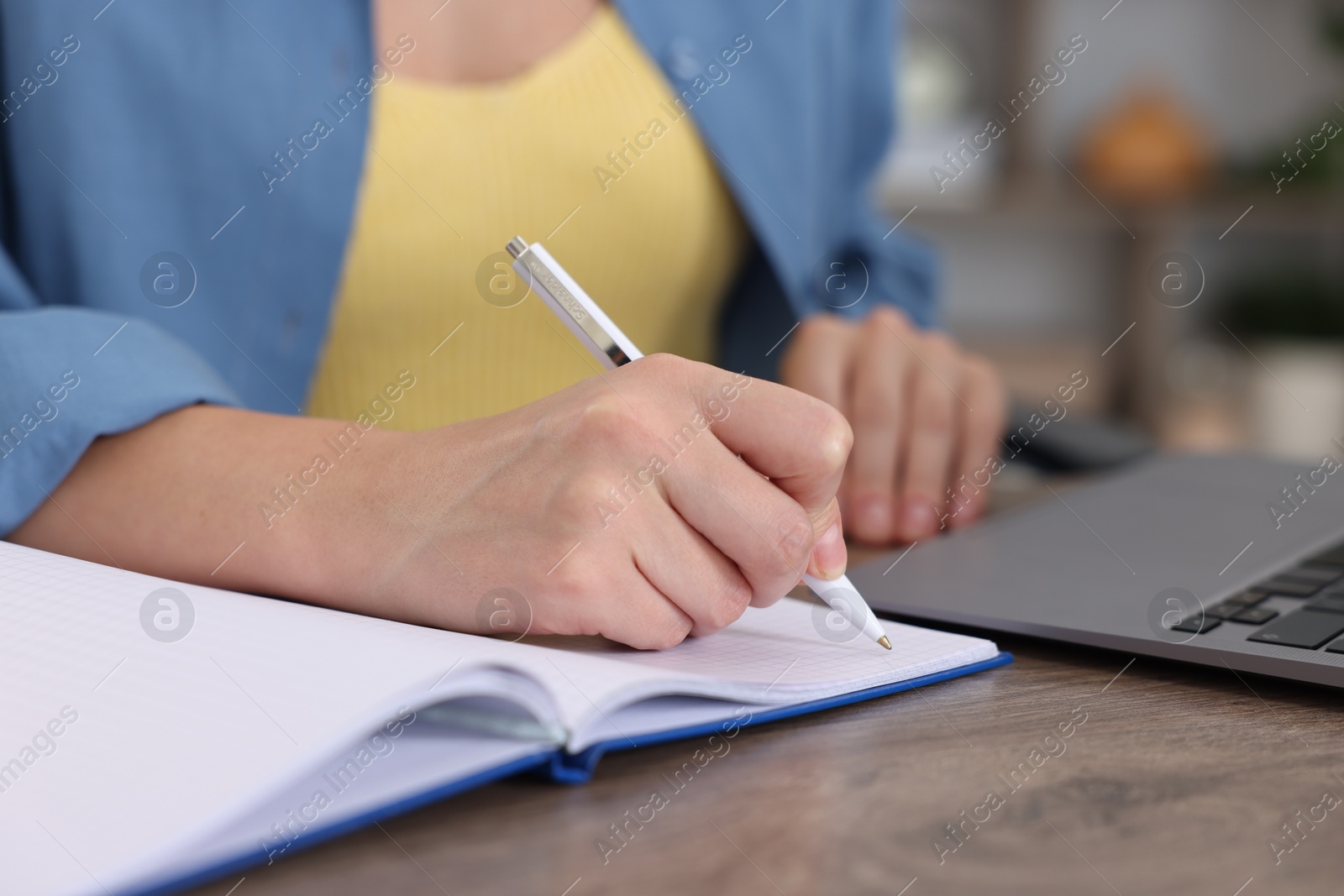 Photo of Woman taking notes while passing online test on laptop at wooden table indoors, closeup