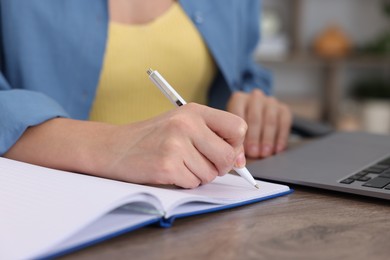 Photo of Woman taking notes while passing online test on laptop at wooden table indoors, closeup