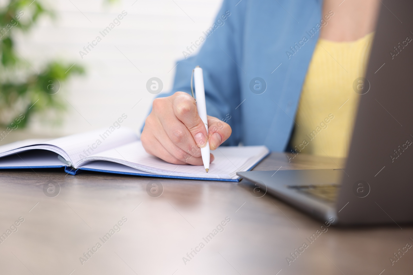 Photo of Woman taking notes while passing online test on laptop at wooden table indoors, closeup