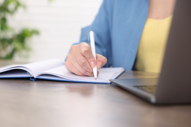 Photo of Woman taking notes while passing online test on laptop at wooden table indoors, closeup