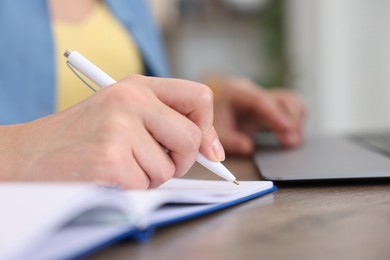 Photo of Woman taking notes while passing online test on laptop at wooden table indoors, closeup