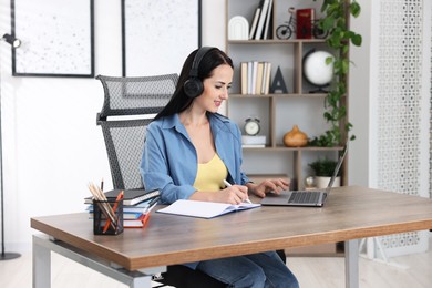 Photo of Woman in headphones taking notes while passing online test on laptop at table indoors