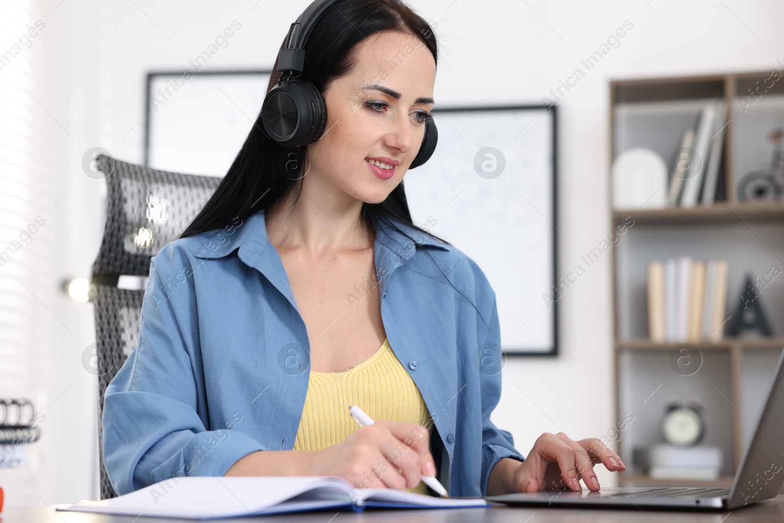 Woman in headphones taking notes while passing online test on laptop at table indoors Photo of Woman in headphones taking notes while passing online test on laptop at table indoors