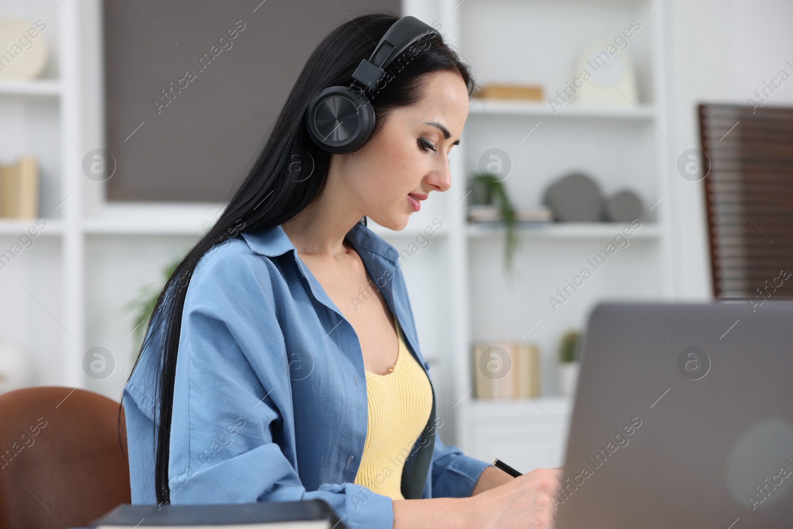 Photo of Woman in headphones taking notes while passing online test via laptop indoors