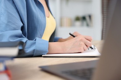 Photo of Woman taking notes while passing online test via laptop at wooden table indoors, closeup