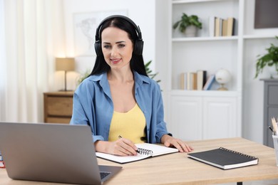 Photo of Woman in headphones taking notes while passing online test via laptop at table indoors
