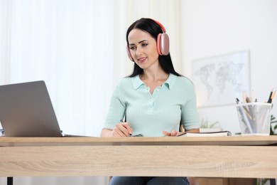 Photo of Woman in headphones taking notes while passing online test via laptop at table indoors