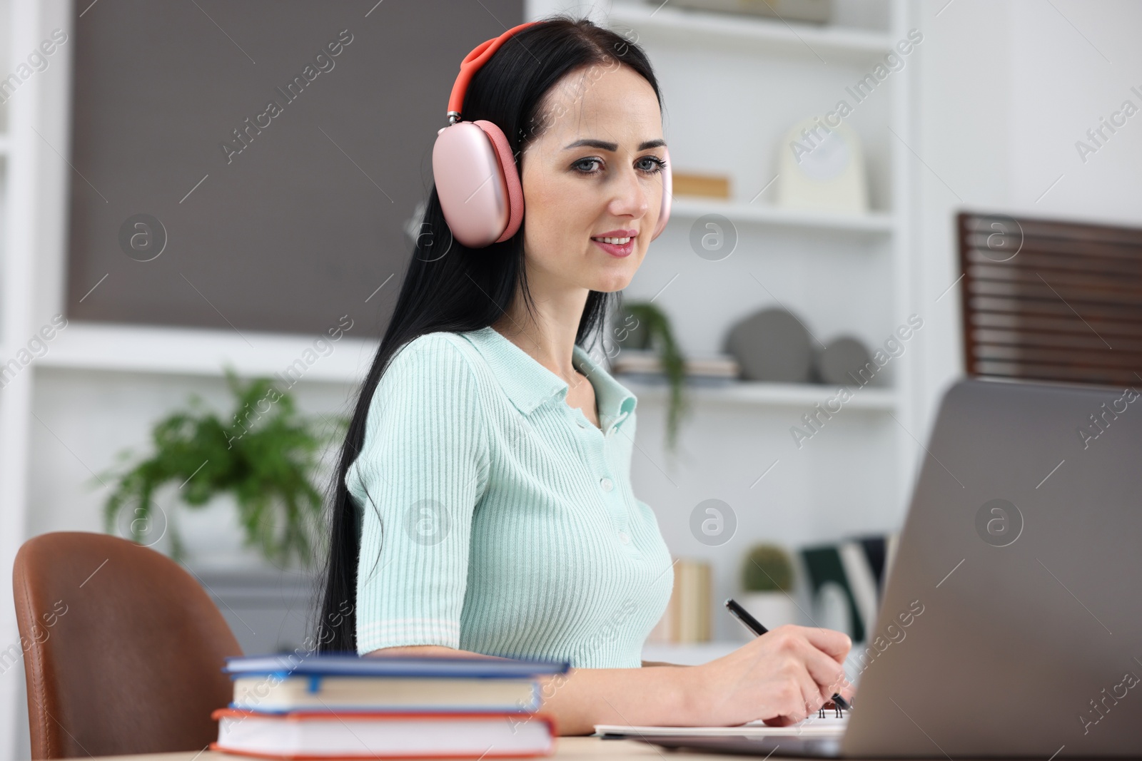 Photo of Woman in headphones taking notes while passing online test via laptop at table indoors