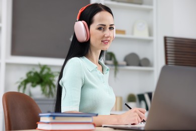 Photo of Woman in headphones taking notes while passing online test via laptop at table indoors